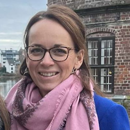 Kvinde med briller og langt lyst hår iført en lyserød tørklæde og blå jakke, smilende med en bygning af røde mursten og vinduer i baggrunden ved vand.