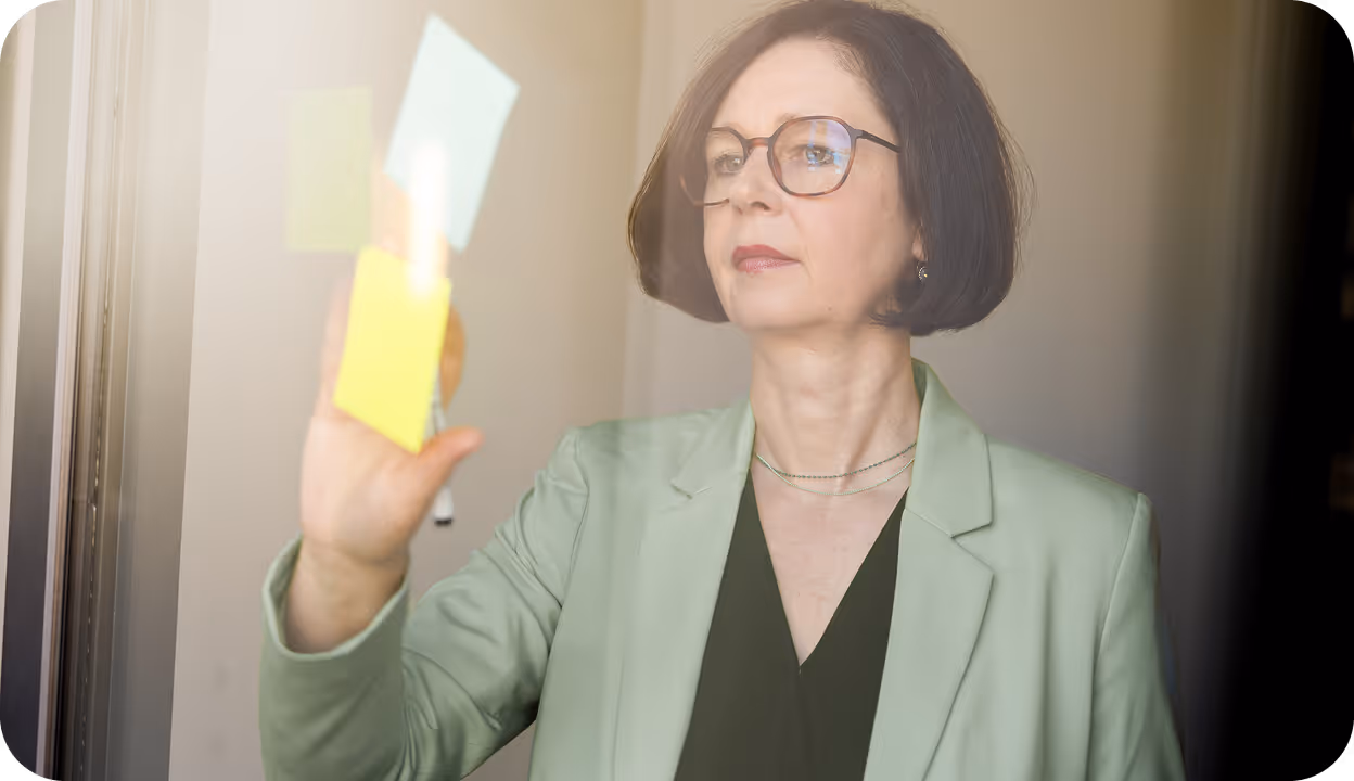 Frau mit Brille ordnet bunte Haftnotizen an einer Glaswand, trägt grünen Blazer.