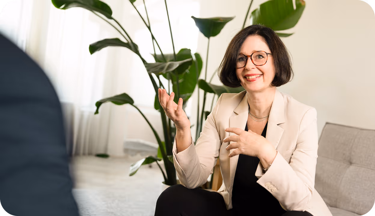 Frau mit Brille und heller Jacke spricht gestikulierend vor einer Pflanze im hellen Raum.