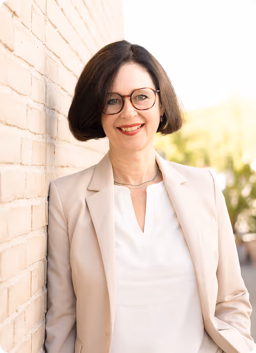 Frau mit Brille und kurzen dunklen Haaren lehnt lächelnd an einer hellen Ziegelwand.