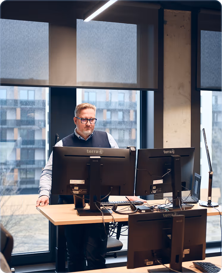 Man wearing glasses standing at desk with three computer monitors in a modern office with large windows.
