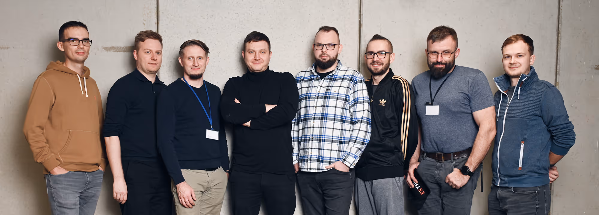 Group of eight casually dressed men standing in a row against a plain concrete wall.