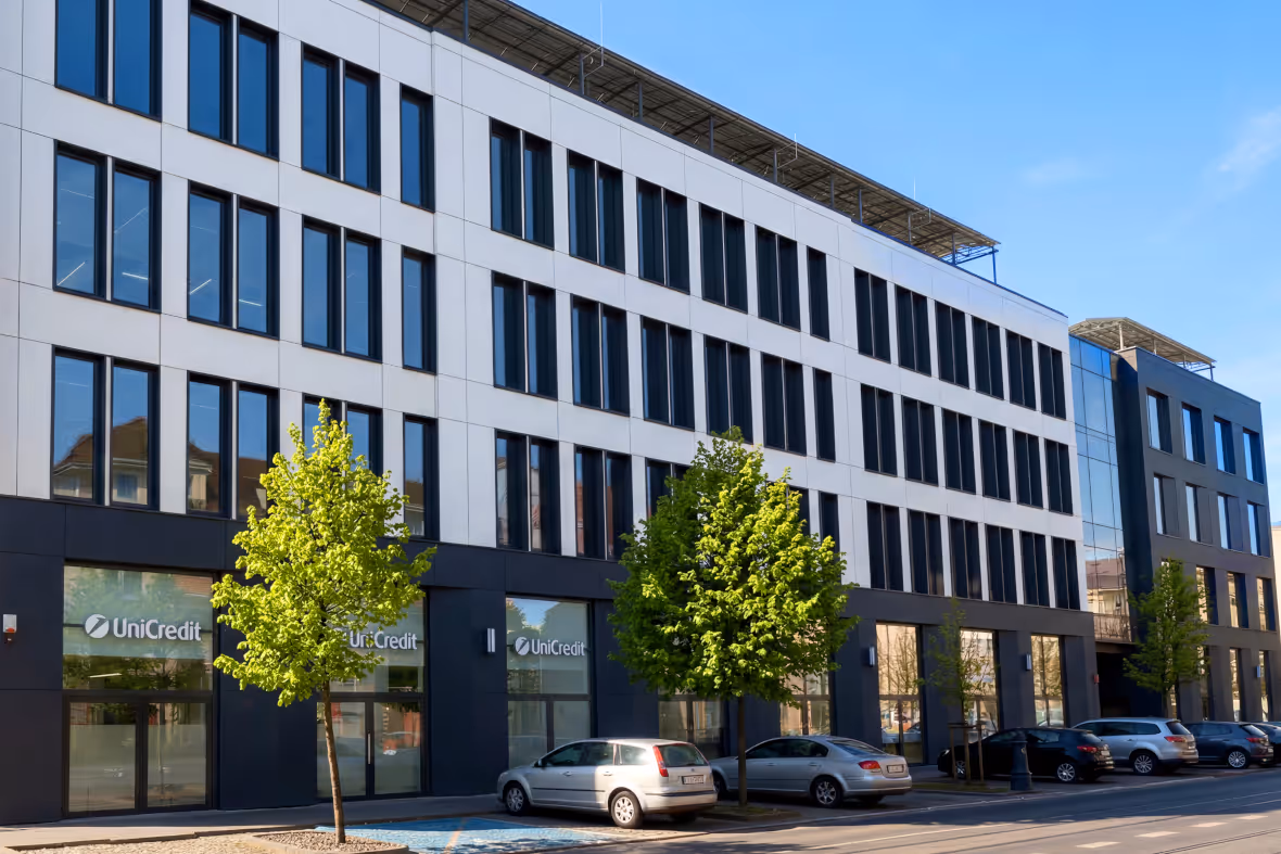 Modern office building with UniCredit signs, multiple windows, and cars parked in front under clear blue sky.