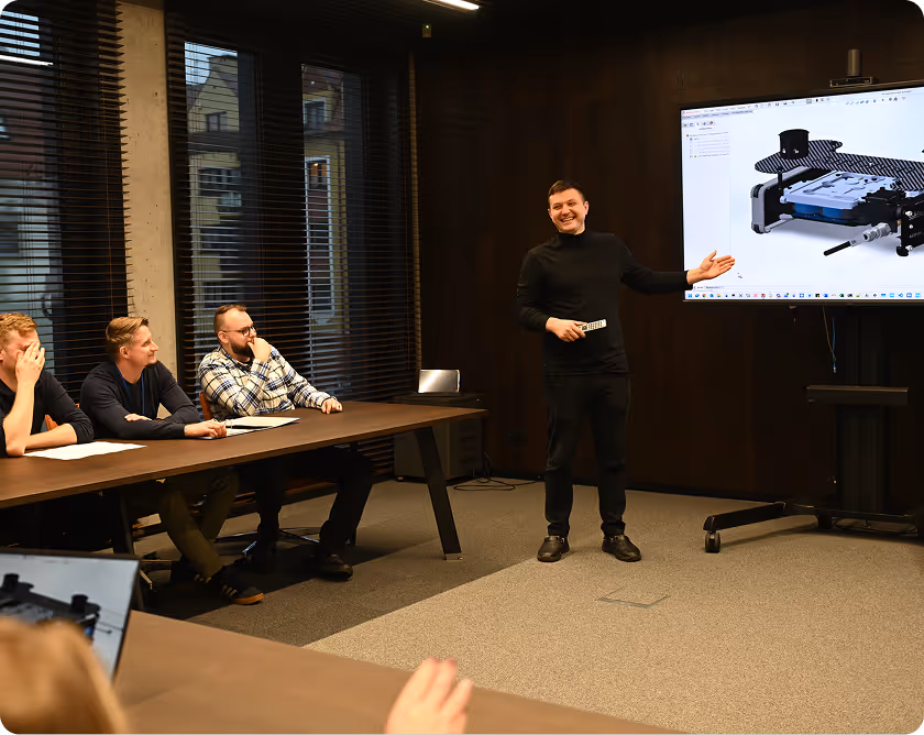 Man in black turtleneck presenting a 3D model on a screen to a group seated at a long table in a meeting room.