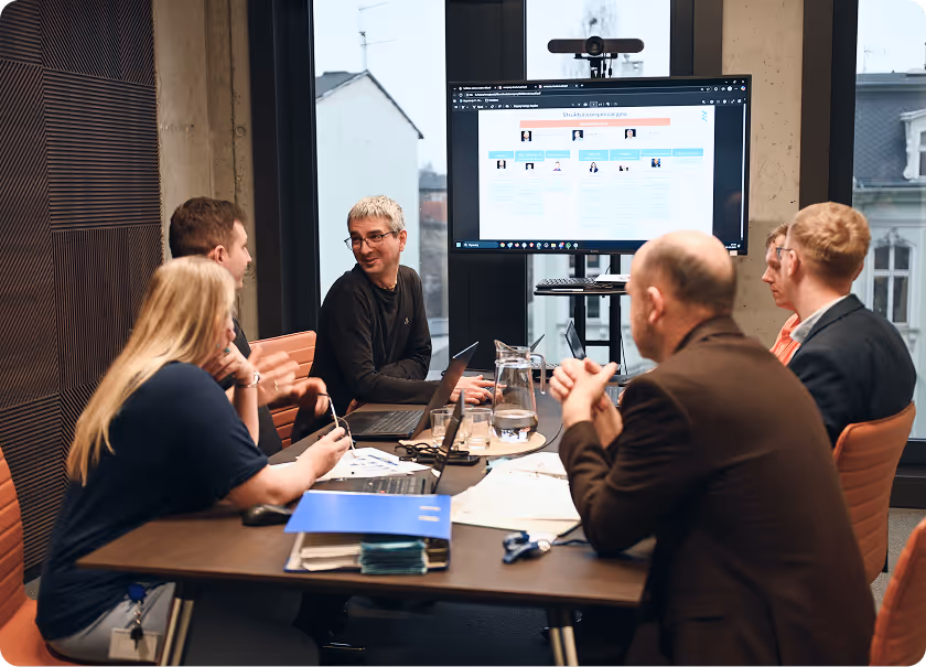 Six people engaged in a meeting around a conference table with a large screen displaying an organizational chart in a modern office.