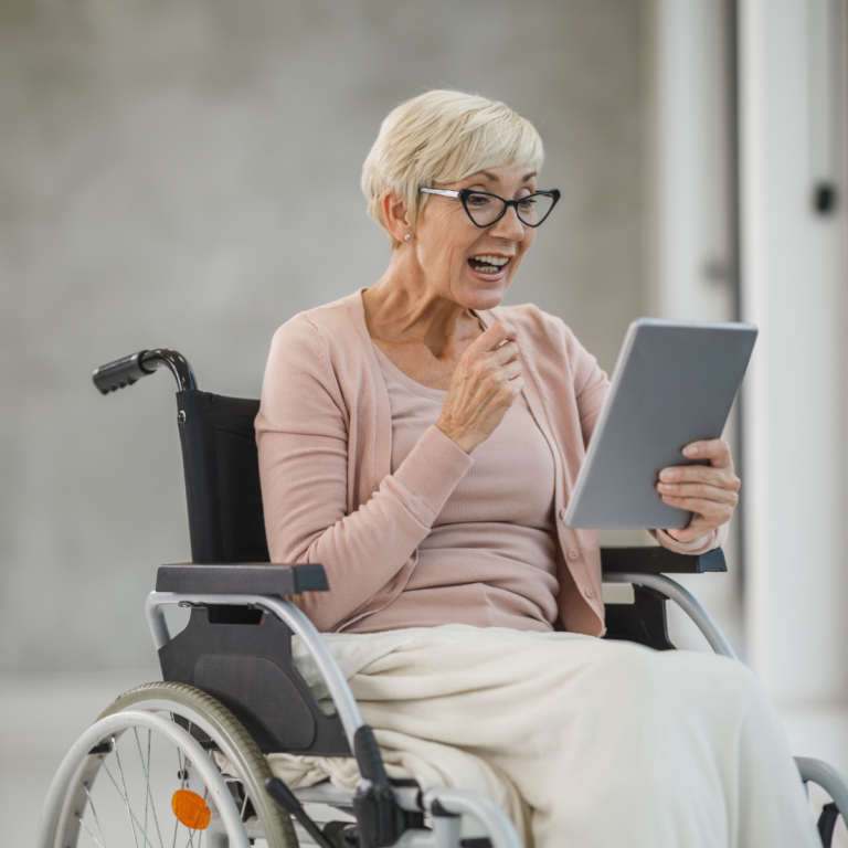 Elderly woman in a wheelchair smiling and gesturing while looking at a tablet.