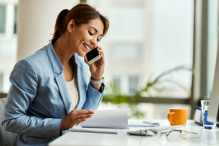 Businesswoman in a blue blazer smiling while talking on her mobile phone and reviewing notes at her office desk.