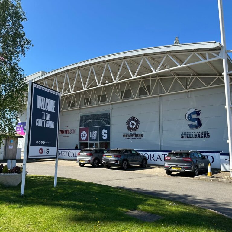 Exterior view of the County Ground, home of Northamptonshire County Cricket Club and the Northamptonshire Steelbacks, with a "Welcome to the County Ground" sign in the foreground and cars parked outside the stadium.