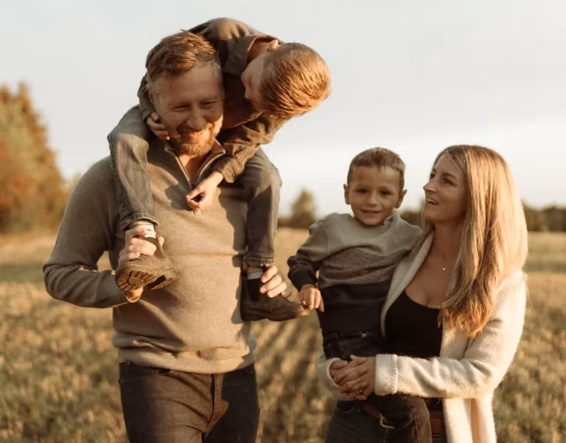 Smiling family of four outdoors: father carries son on shoulders, mother holds younger son.