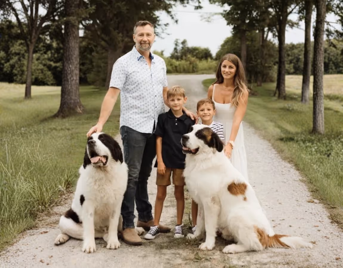 Family of four posing outdoors on a tree-lined path with two large Saint Bernard dogs.