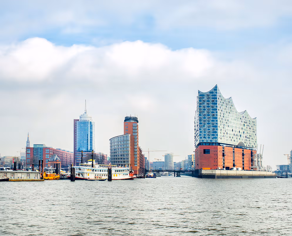 View of the Elbphilharmonie concert hall and surrounding buildings along the waterfront in Hamburg, Germany under a cloudy sky.