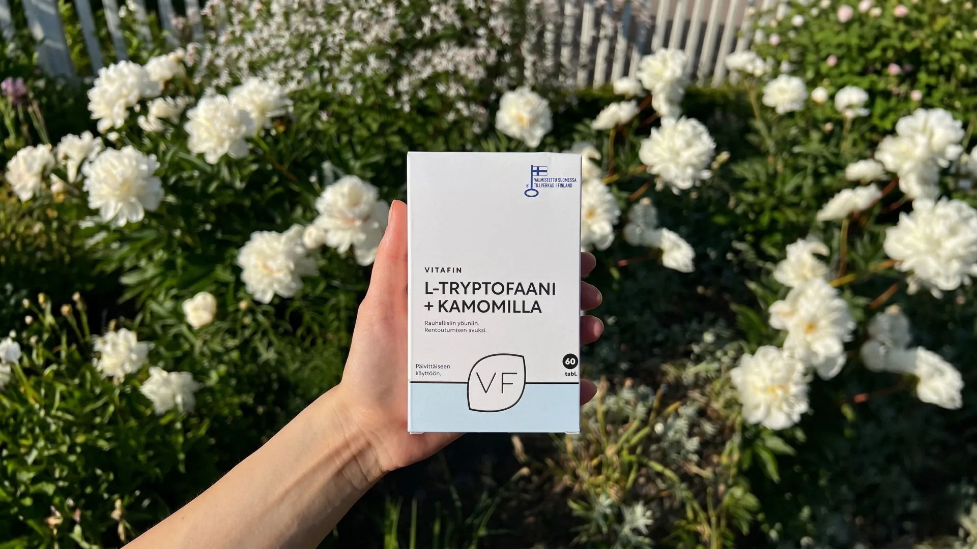 Vitafin L-Tryptofaani + Kamomilla kädessä kukkivien pioneiden edessä.