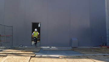 Ouvrier de construction portant un casque et un gilet fluorescent marche vers une porte d'un mur gris.