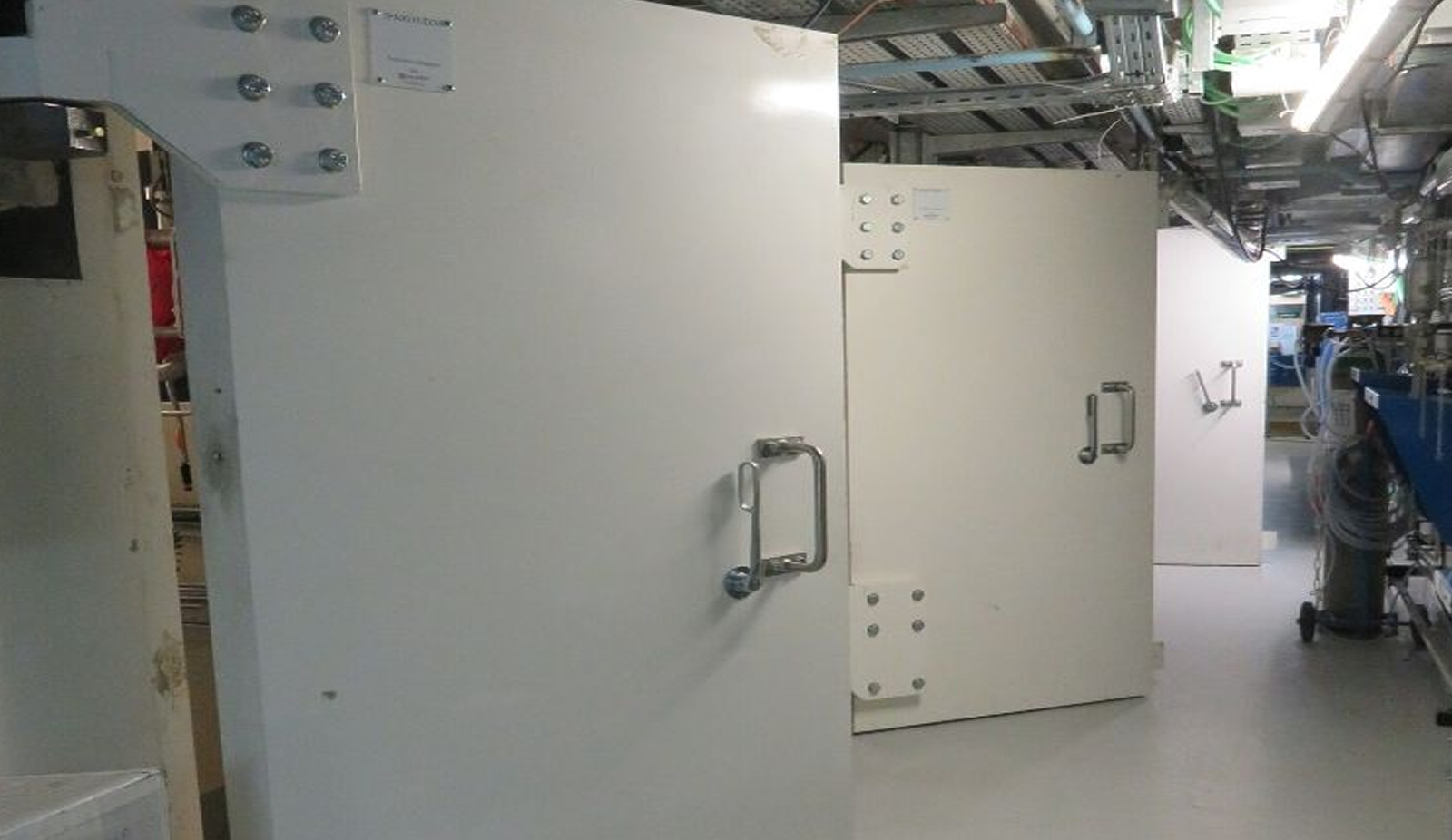 Couloir industriel avec grandes portes blanches renforcées et équipements mécaniques sur le côté droit.
