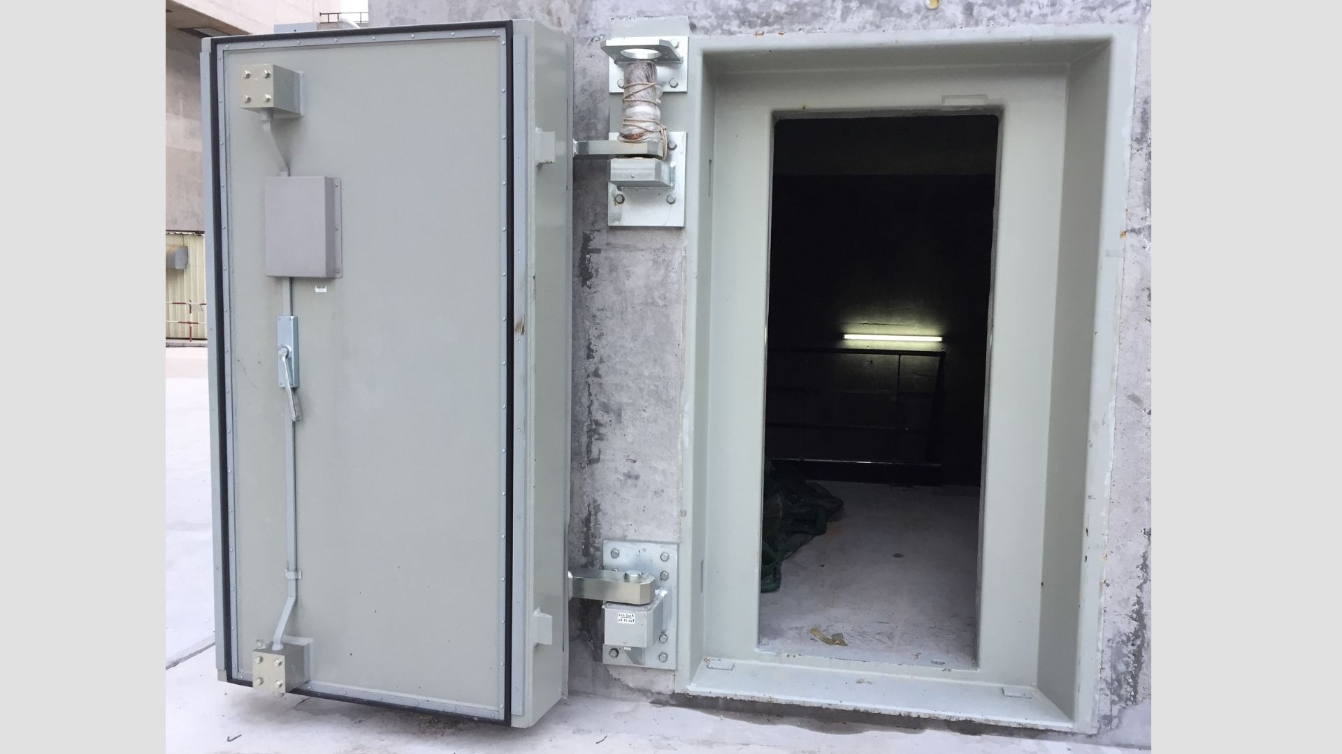 Porte blindée industrielle grise ouverte avec intérieur sombre et éclairage fluorescent au fond.