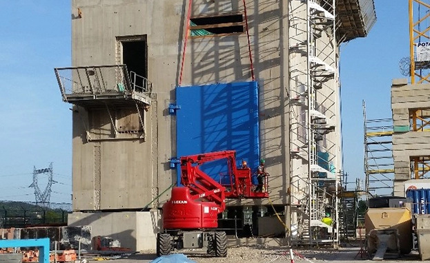 Construction site with concrete building, red machinery, scaffolding, and workers lifting a blue panel.