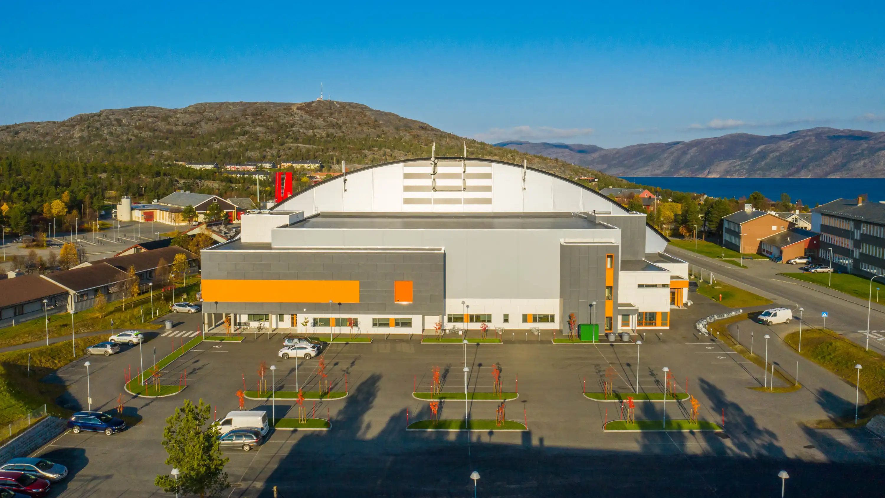 Moderne flerbrukshall med grå og oransje fasade, parkeringsplass og fjell i bakgrunnen.