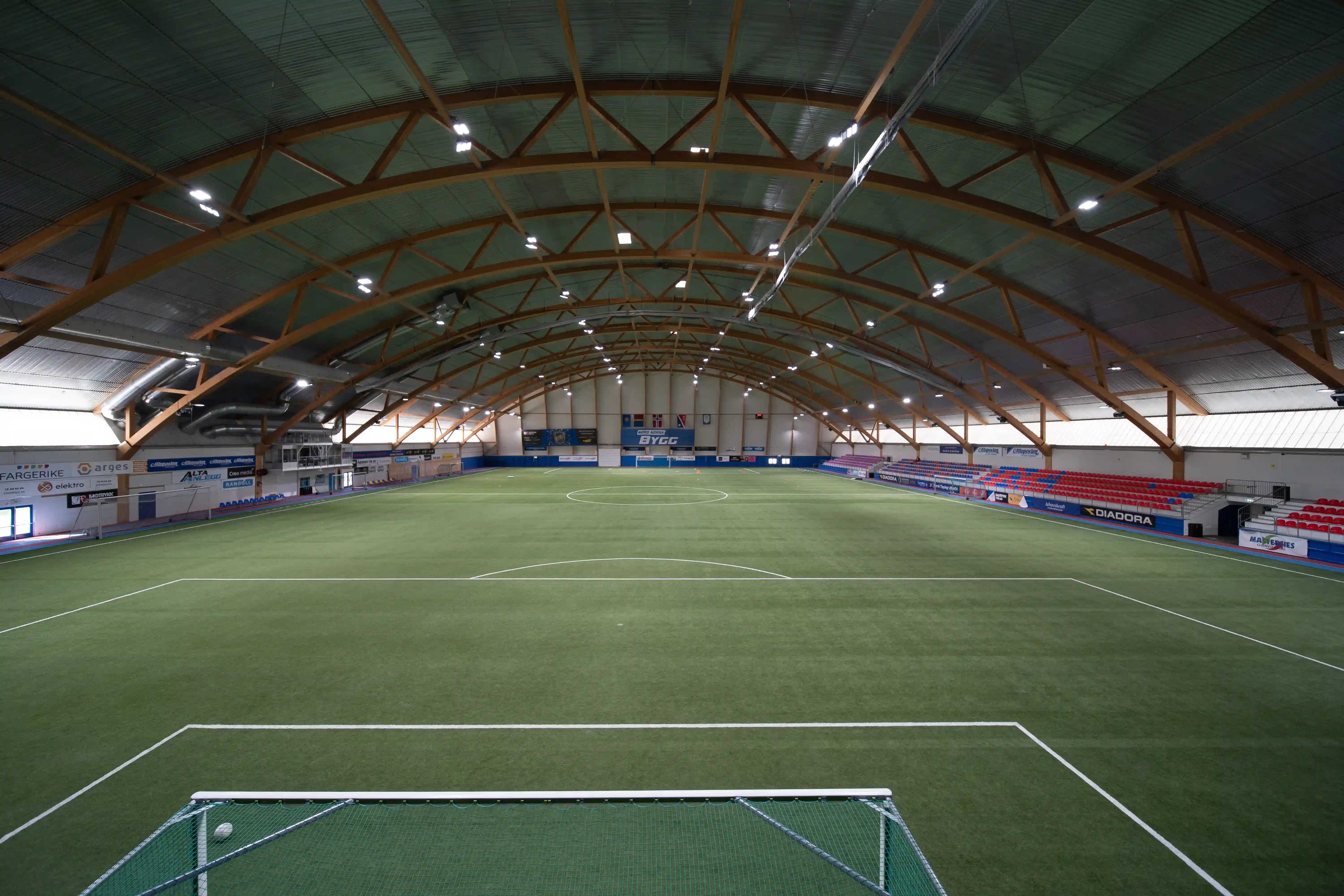 Innendørs fotballbane med kunstgress, mål og tilskuerplasser under en buet takstruktur.