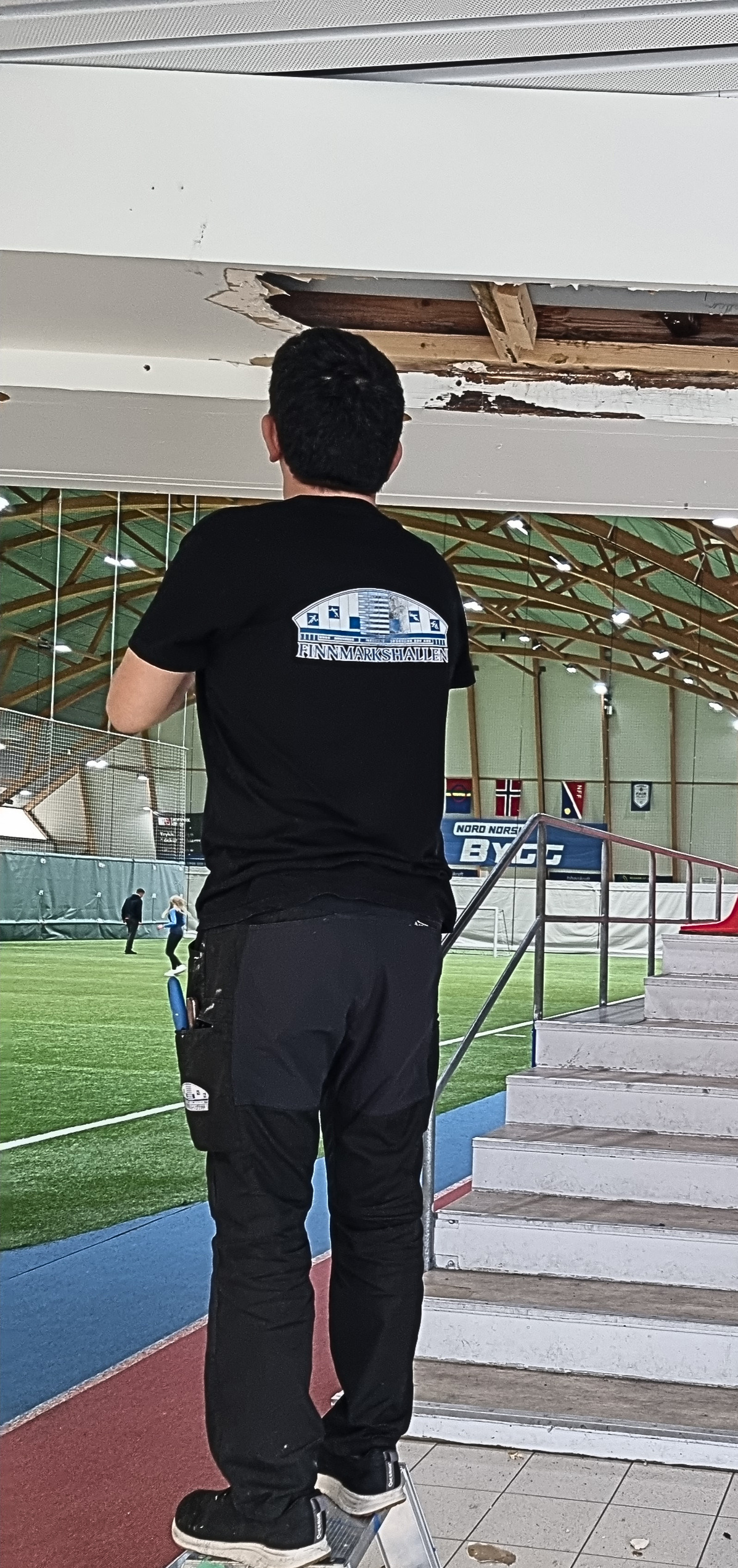 Person står på en krakk ved et innendørs fotballbanebygg med skadet tak.