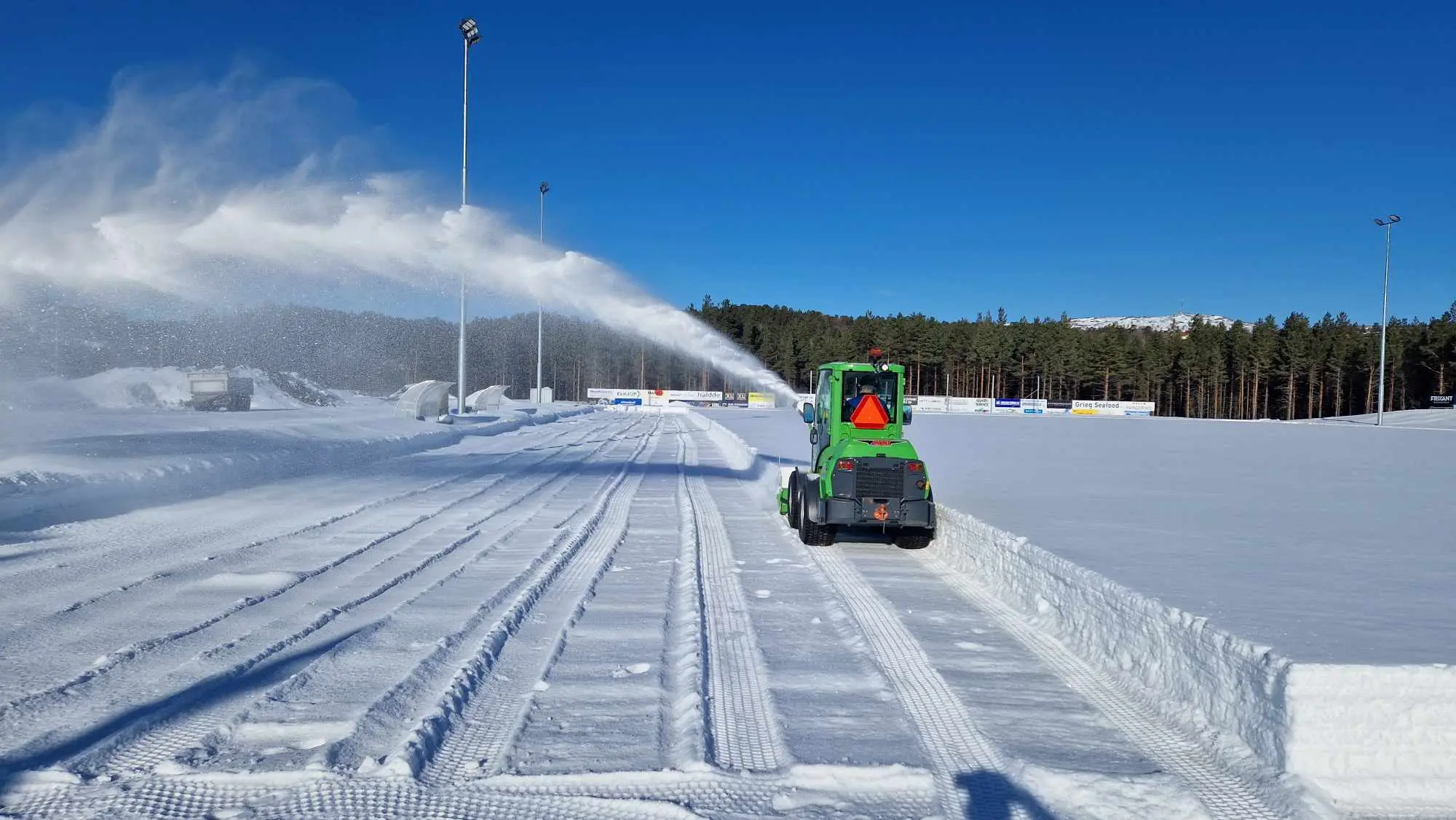 Grønn snøfreser rydder en snødekt idrettsbane under blå himmel.