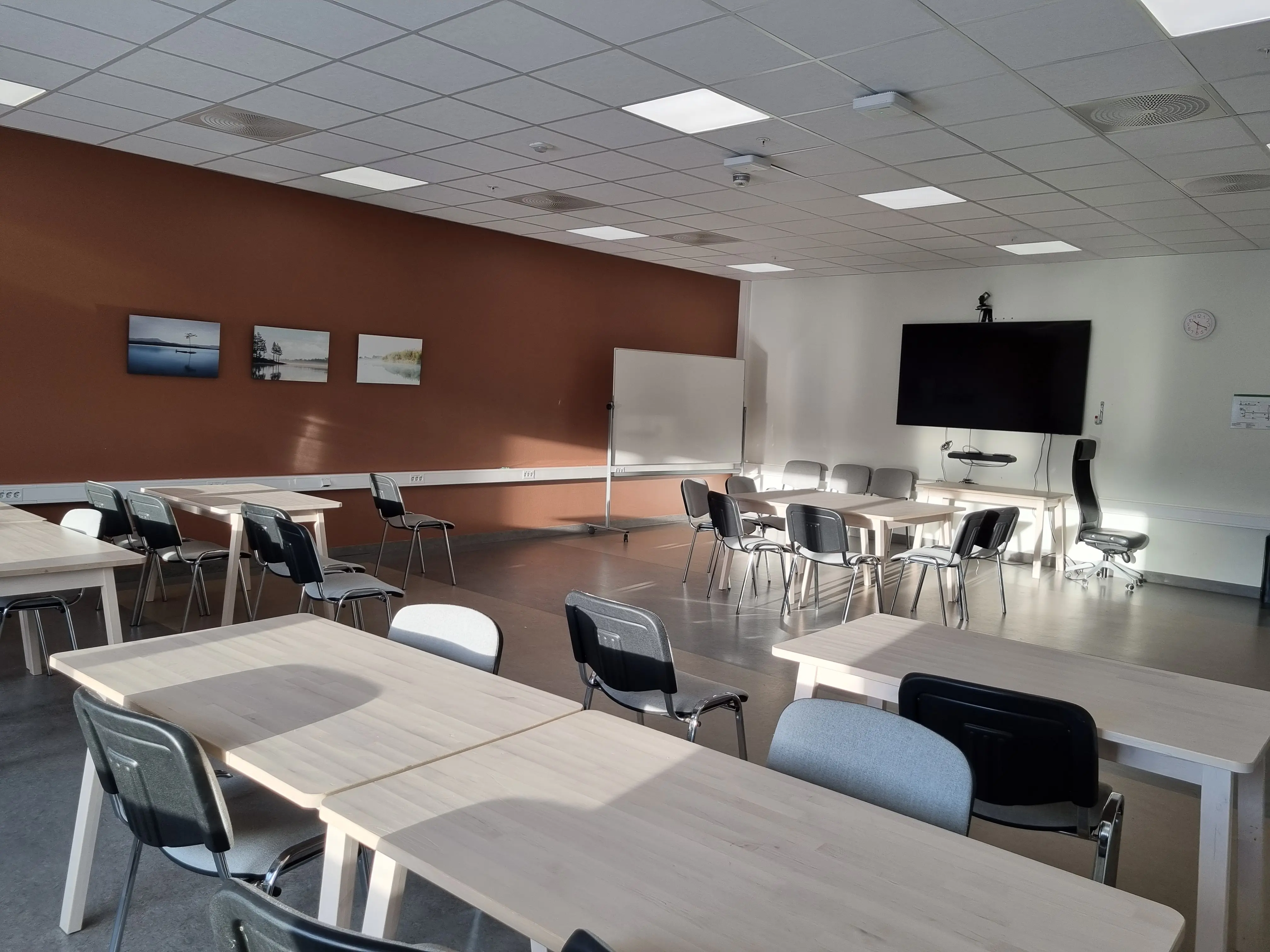 Tomt klasserom med flere bord, stoler, whiteboard, TV-skjerm og bilder på veggen.