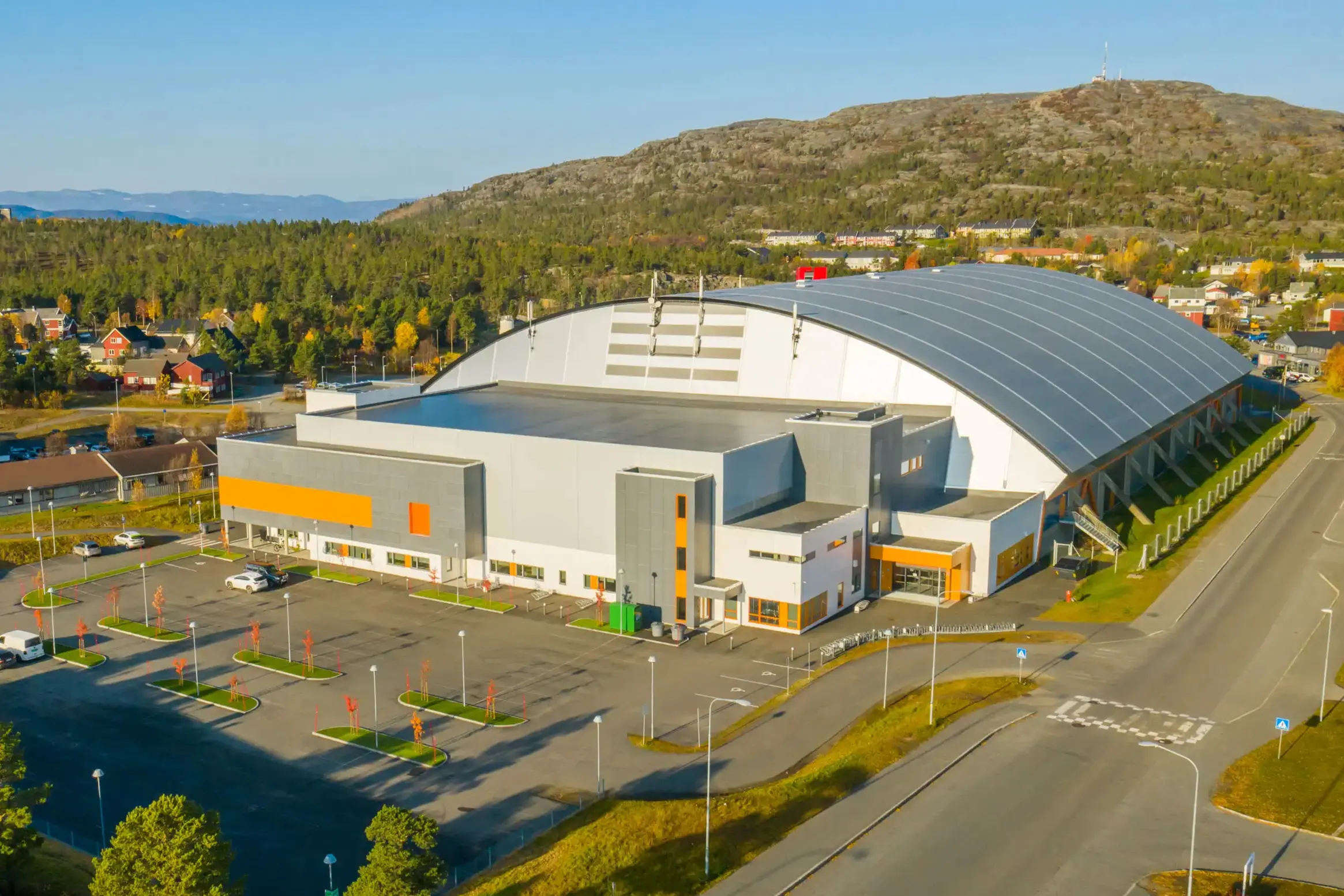 Moderne idrettshall med stor buet tak, parkeringsplass og fjell og skog i bakgrunnen.