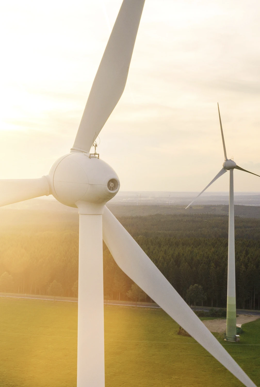 Éoliennes blanches dans un champ vert au coucher du soleil avec une forêt en arrière-plan.