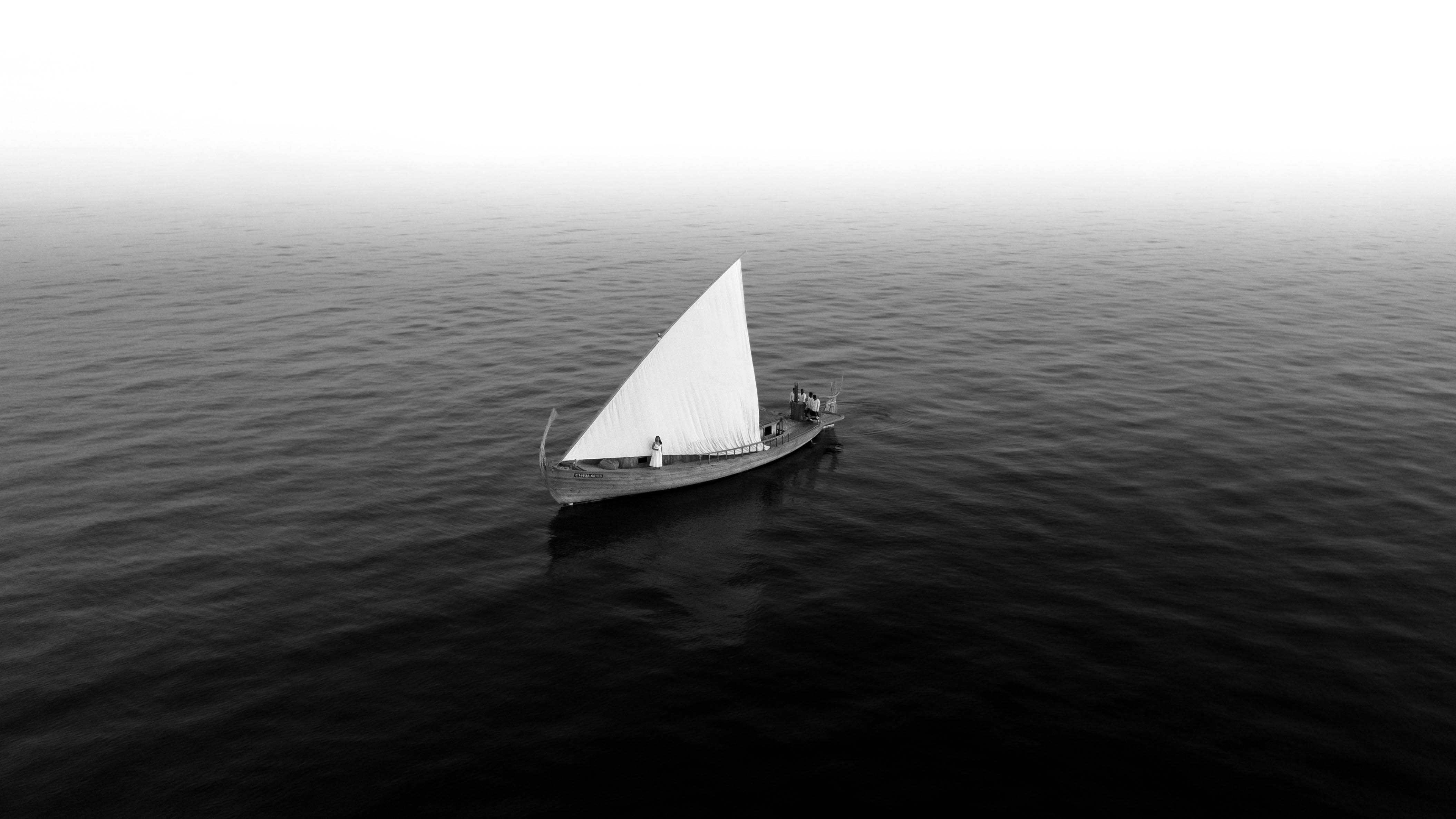 Small sailboat with a single triangular white sail on calm water with three people aboard.