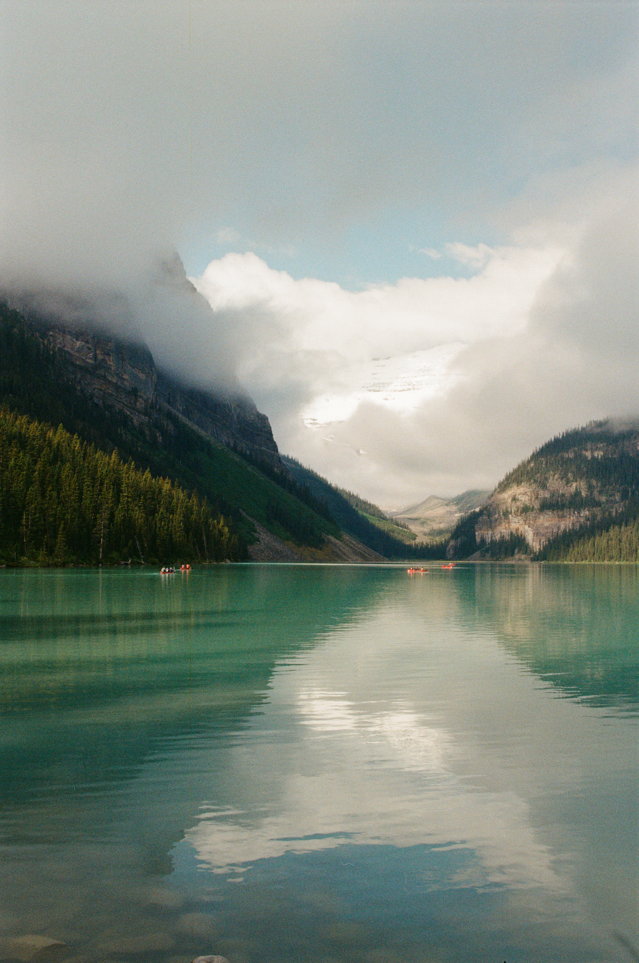 Calm turquoise lake with red canoes, surrounded by pine forests and misty mountains under clouds.
