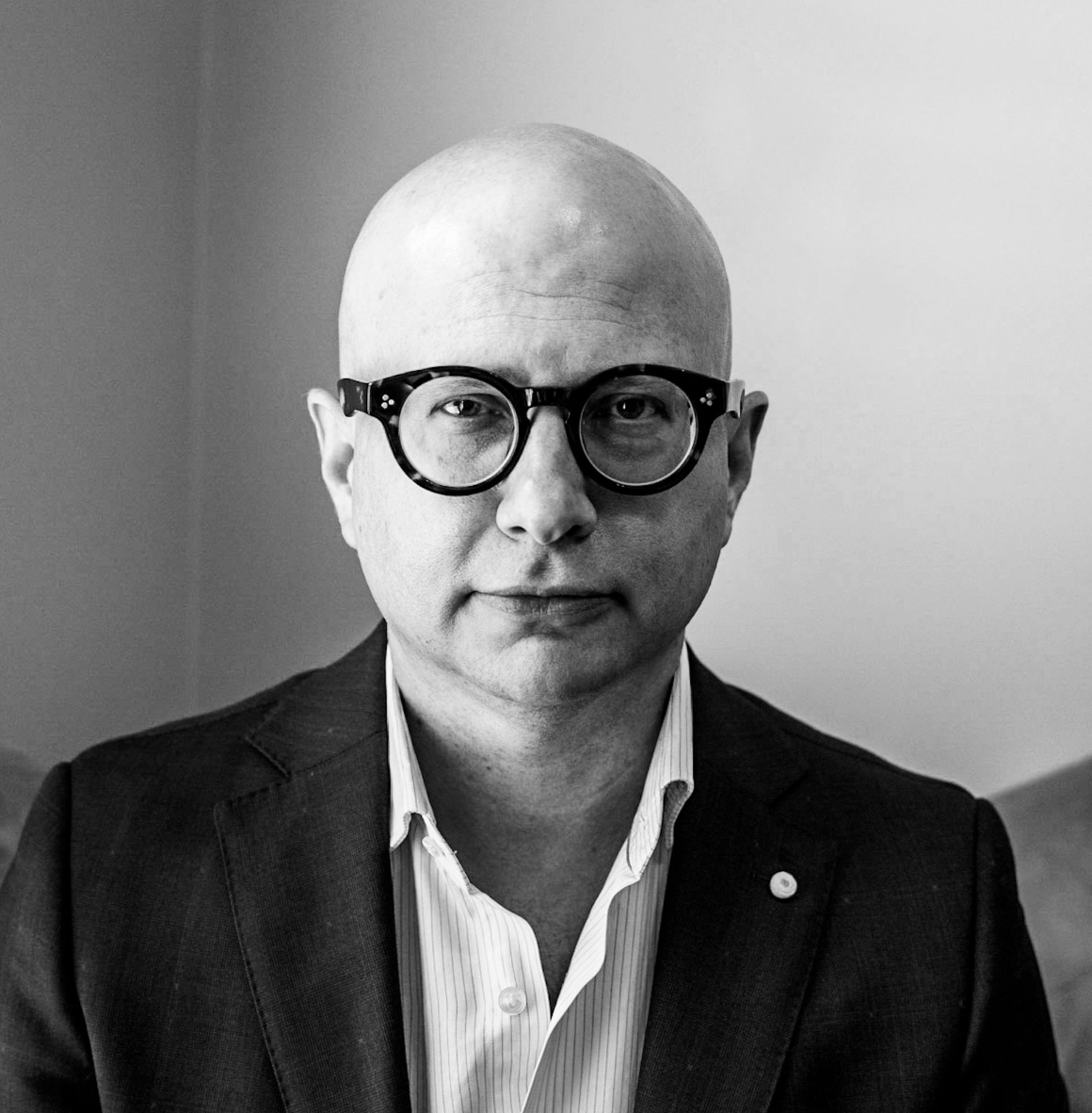 Headshot of a bald man with round dark glasses wearing a suit jacket and striped shirt.