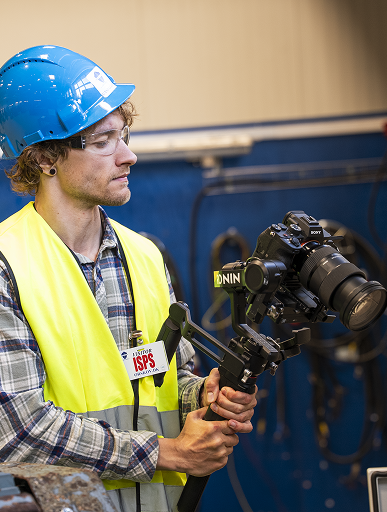 Mand med blå hjelm og gul vest holder kamera med stabilisator i industriel miljø.