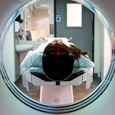 Person lies inside a CT scan machine with red laser cross on their head, in a medical room.
