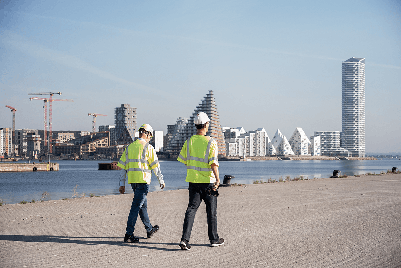 To bygningsarbejdere i gule veste og hjelme går ved havnefronten med moderne bygninger og kraner i baggrunden.