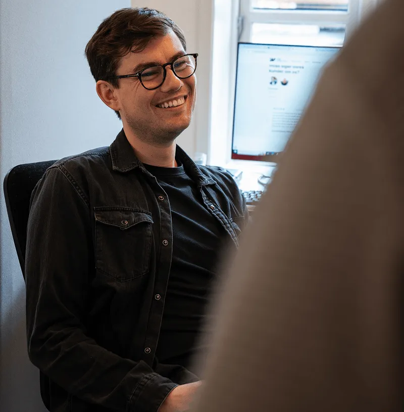 Ung mand med tørklæde og briller smiler, mens han sidder ved et skrivebord med en computerskærm.