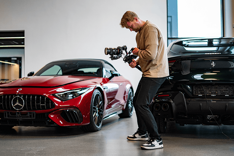 Mand filmer rødt Mercedes sportsvogn ved siden af sort Ferrari i showroom.