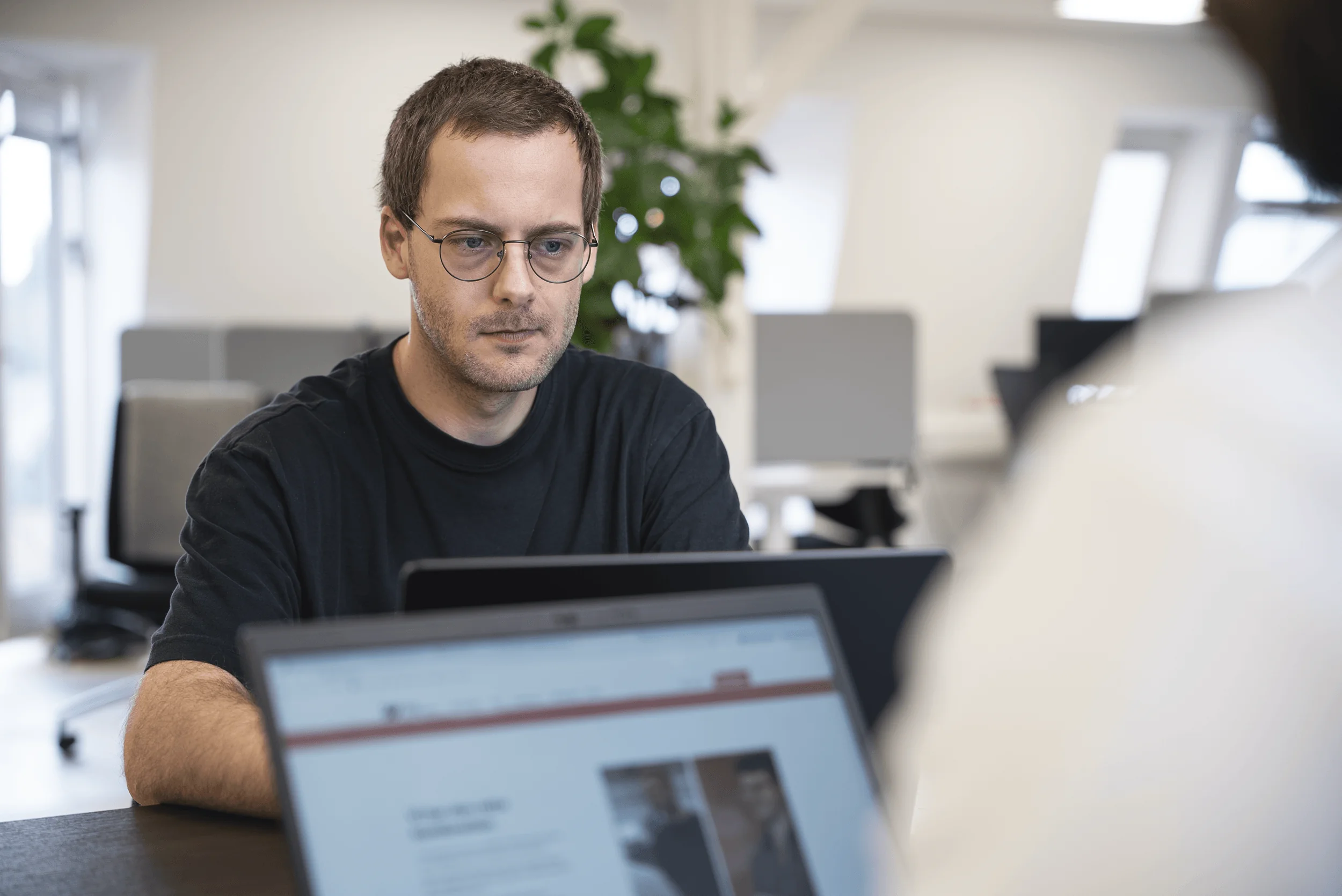 Mand med briller arbejder koncentreret ved bærbar computer i kontormiljø.