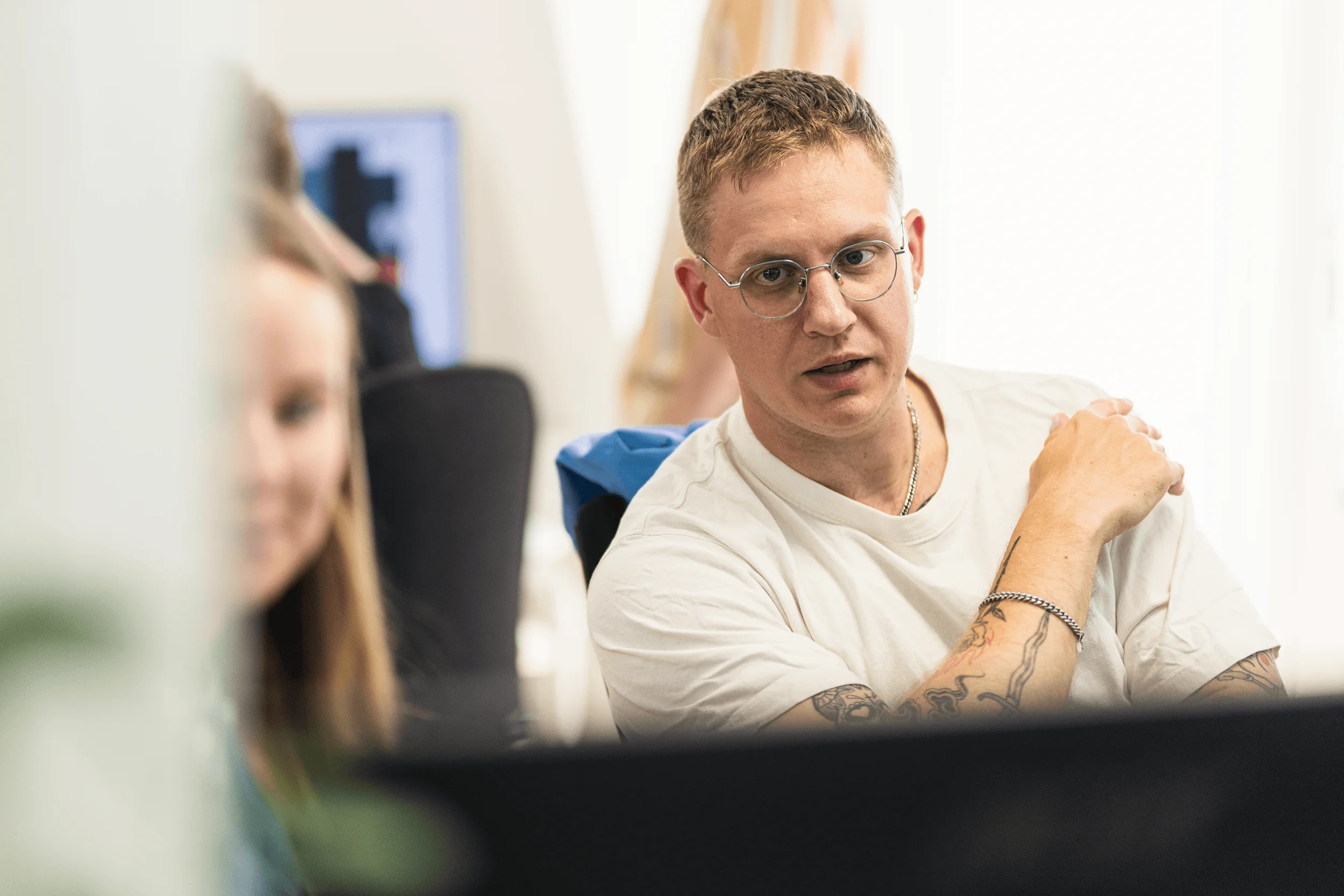 Mand med briller og tatoveringer taler fokuseret til en anden person i et lyst kontormiljø.