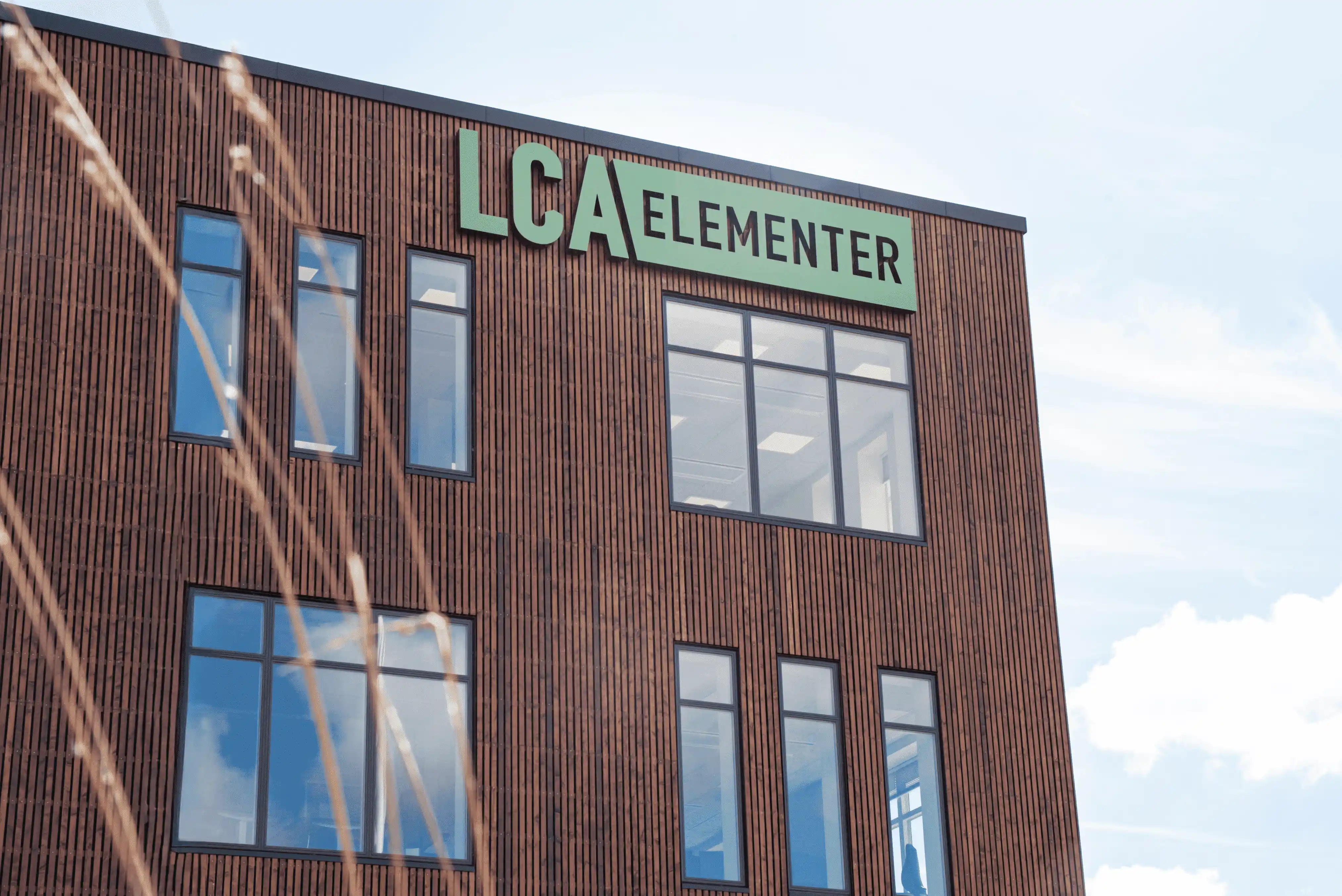 Facade af træbygning med skiltet LCA Elementer og reflekterede skyer i vinduer.