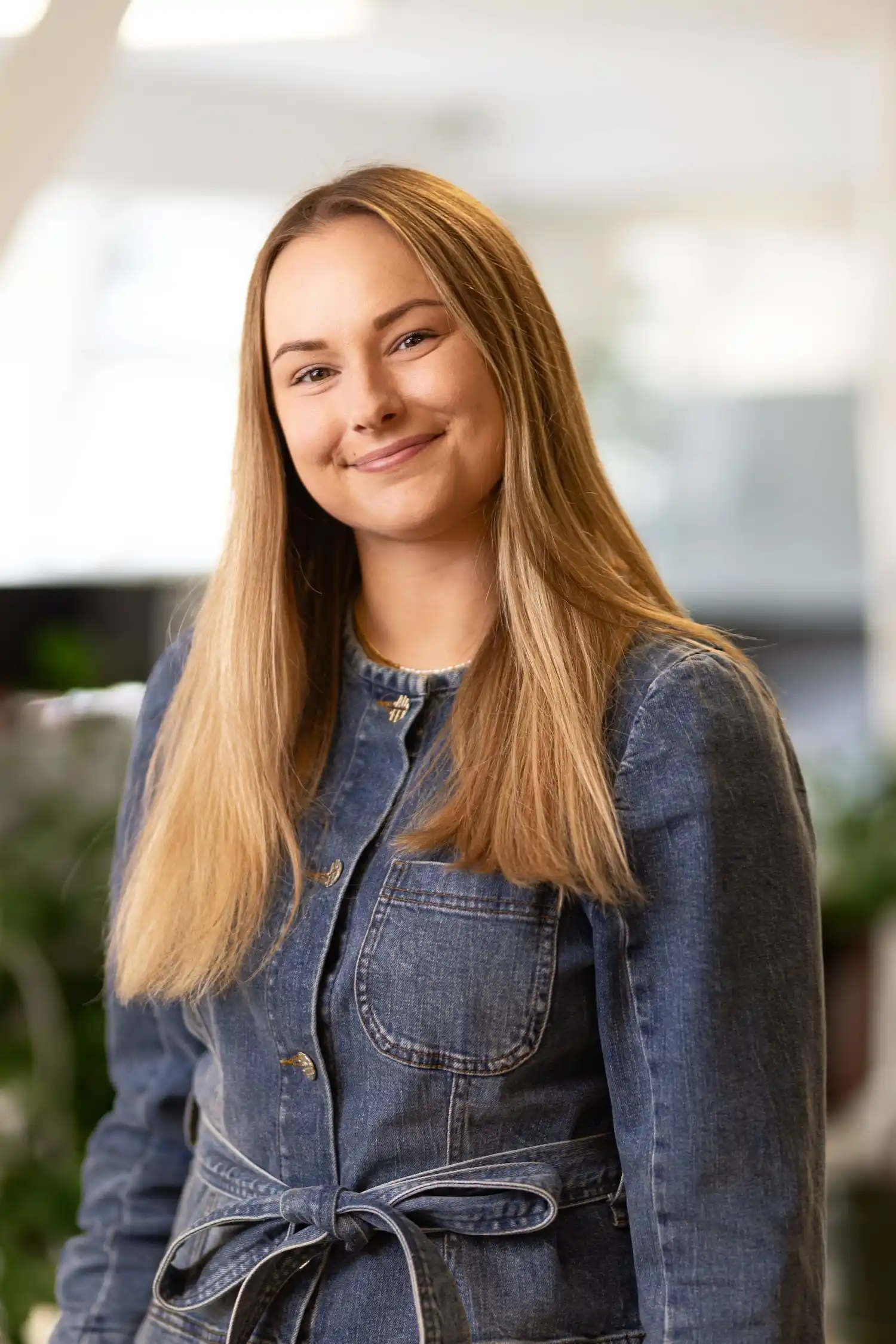 Smilende ung kvinde med langt blondt hår iført en denimjakke med bælte foran.