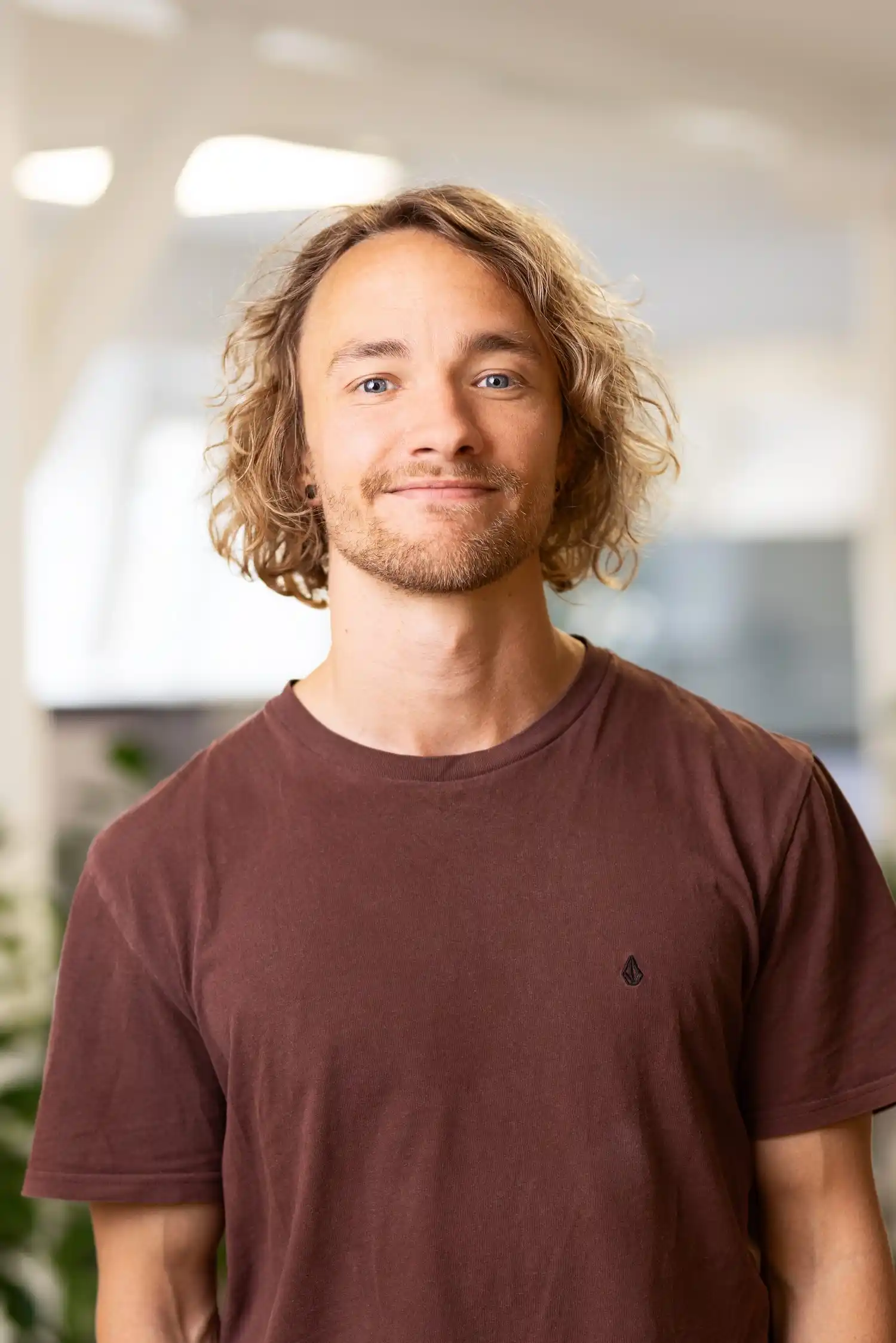 Mand med blondt bølget hår og skæg iført en brun t-shirt smilende i et lyst rum.