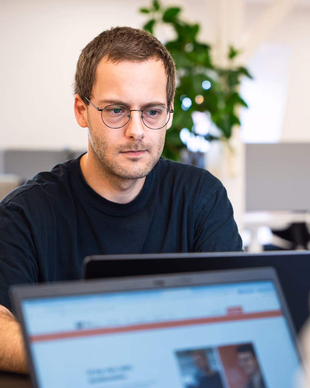 Mand med briller arbejder fokuseret på en bærbar computer i et kontormiljø.