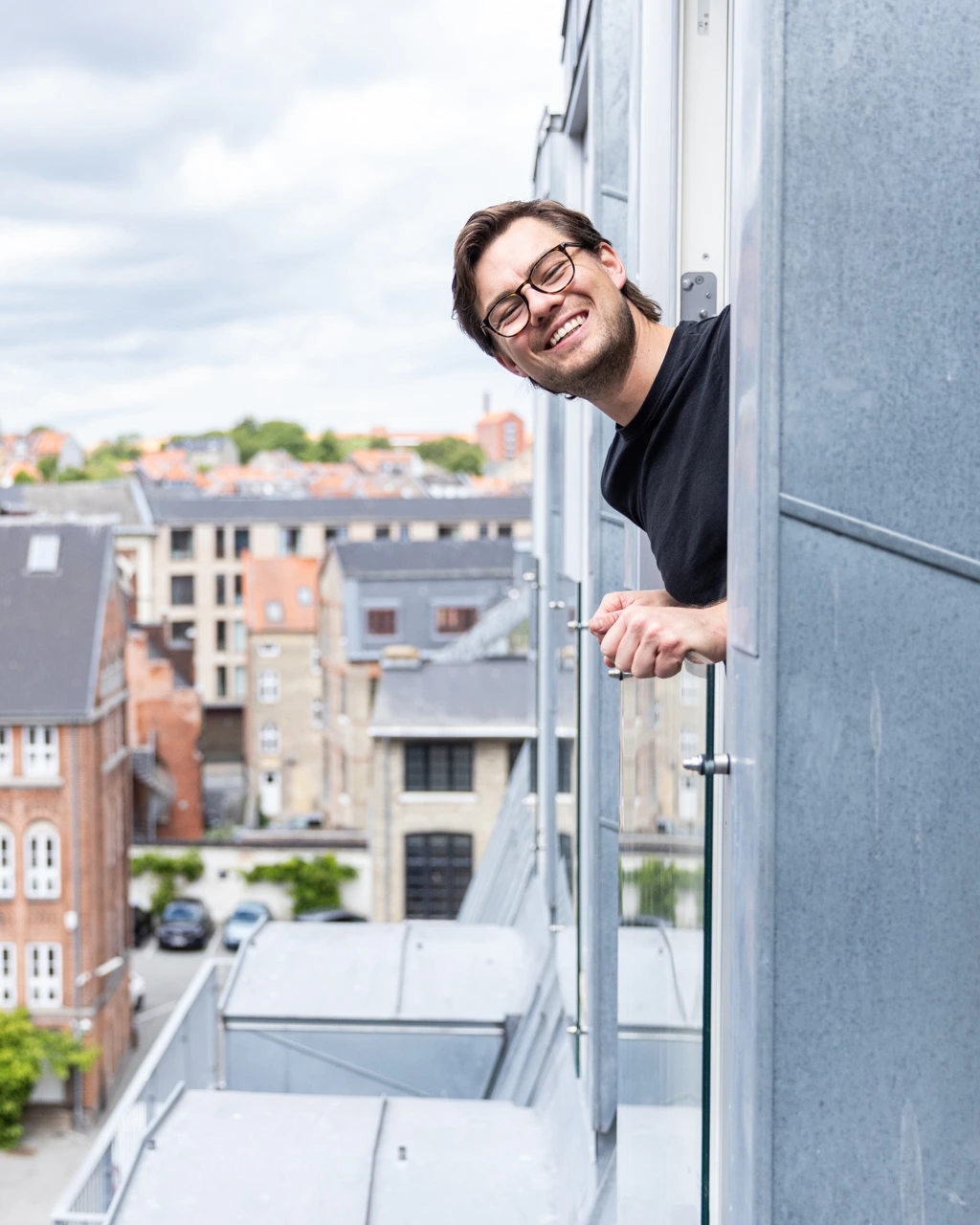 Smilende mand med briller kigger ud fra en balkon i en by med bygninger i baggrunden.