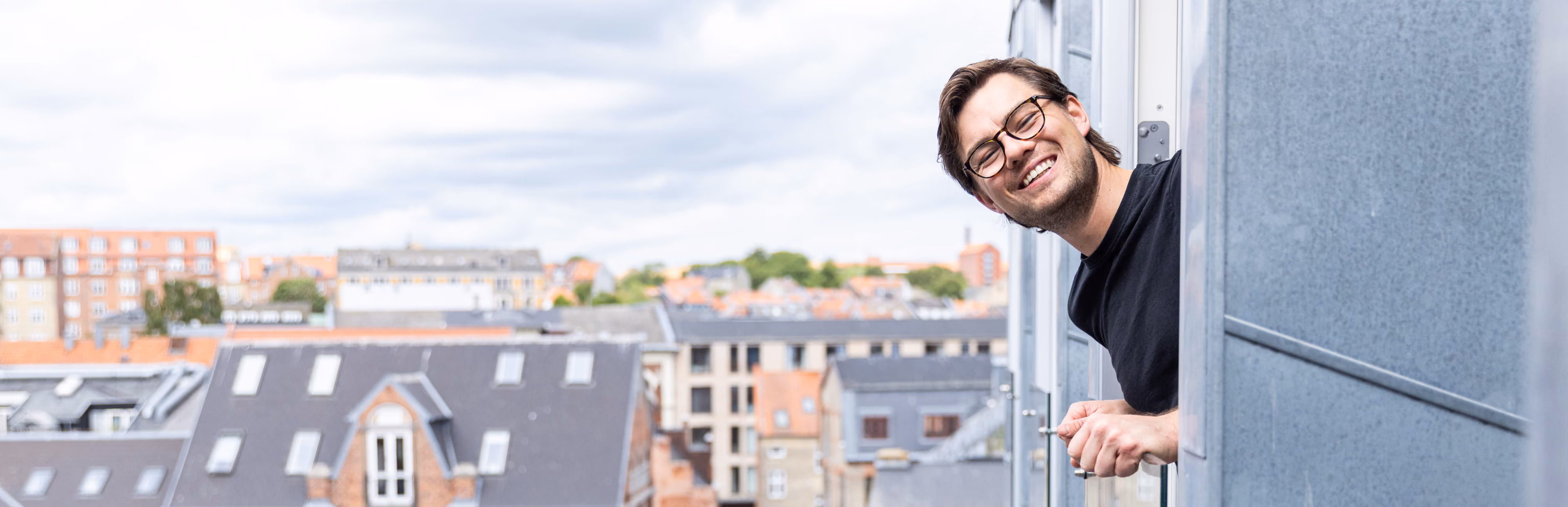 Mand med briller smiler og kigger ud af et vindue mod byens tage.