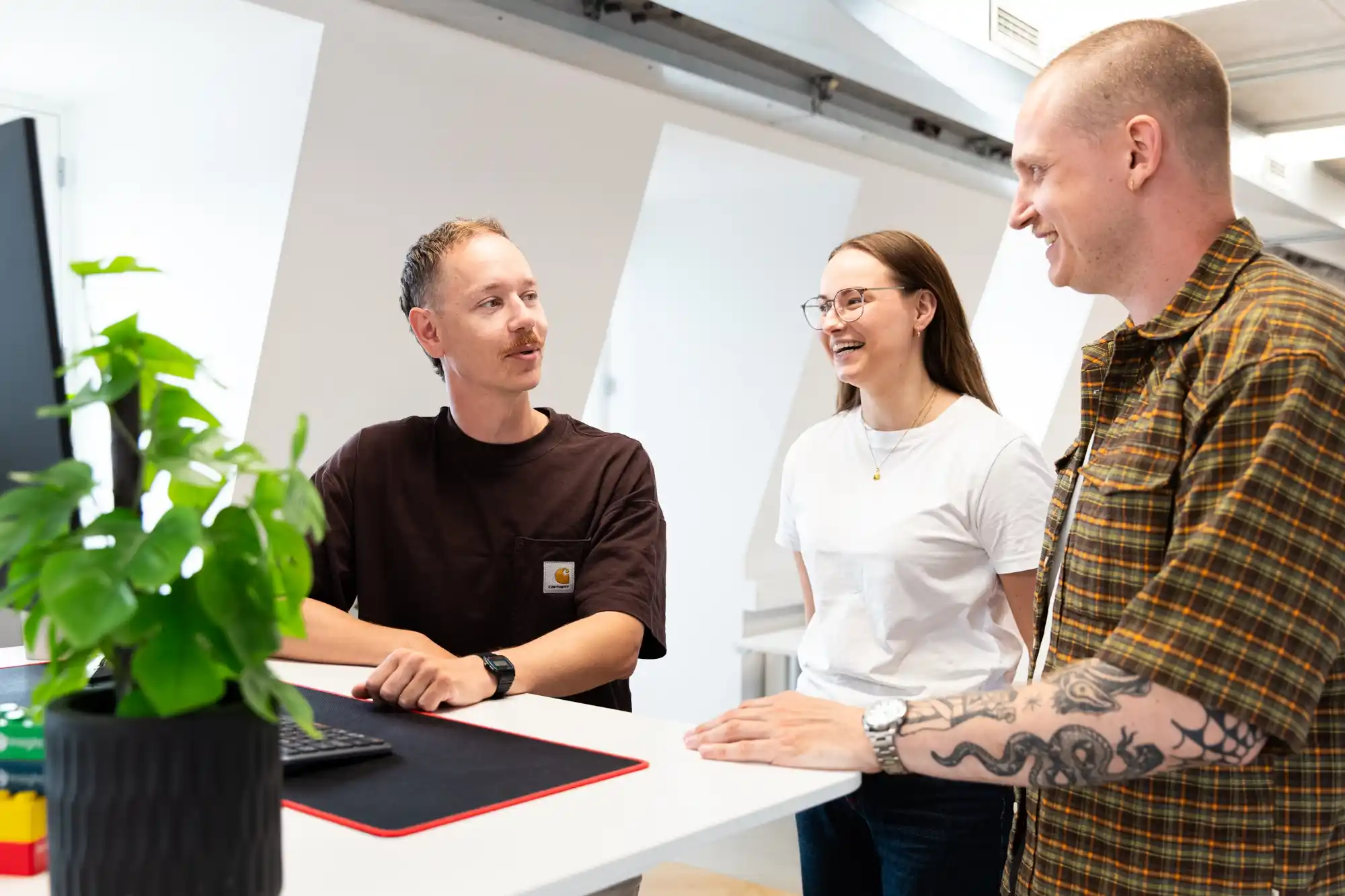 Tre personer smilende og snakkende sammen ved et ståbord i et lyst kontormiljø.