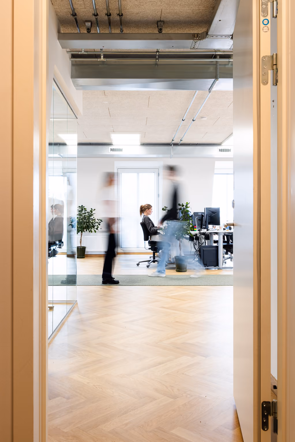 Tos personer sløret i bevægelse i kontor med en siddende kvinde ved skrivebord og planter.
