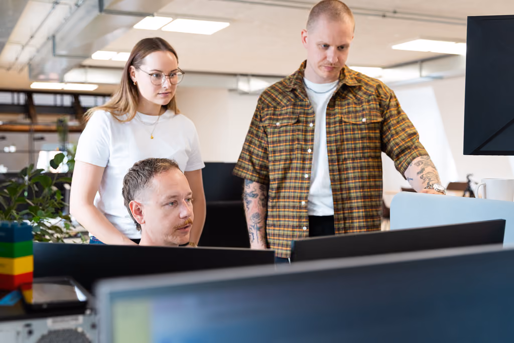 Tre kolleger samarbejder og ser på computerskærm i moderne kontormiljø.