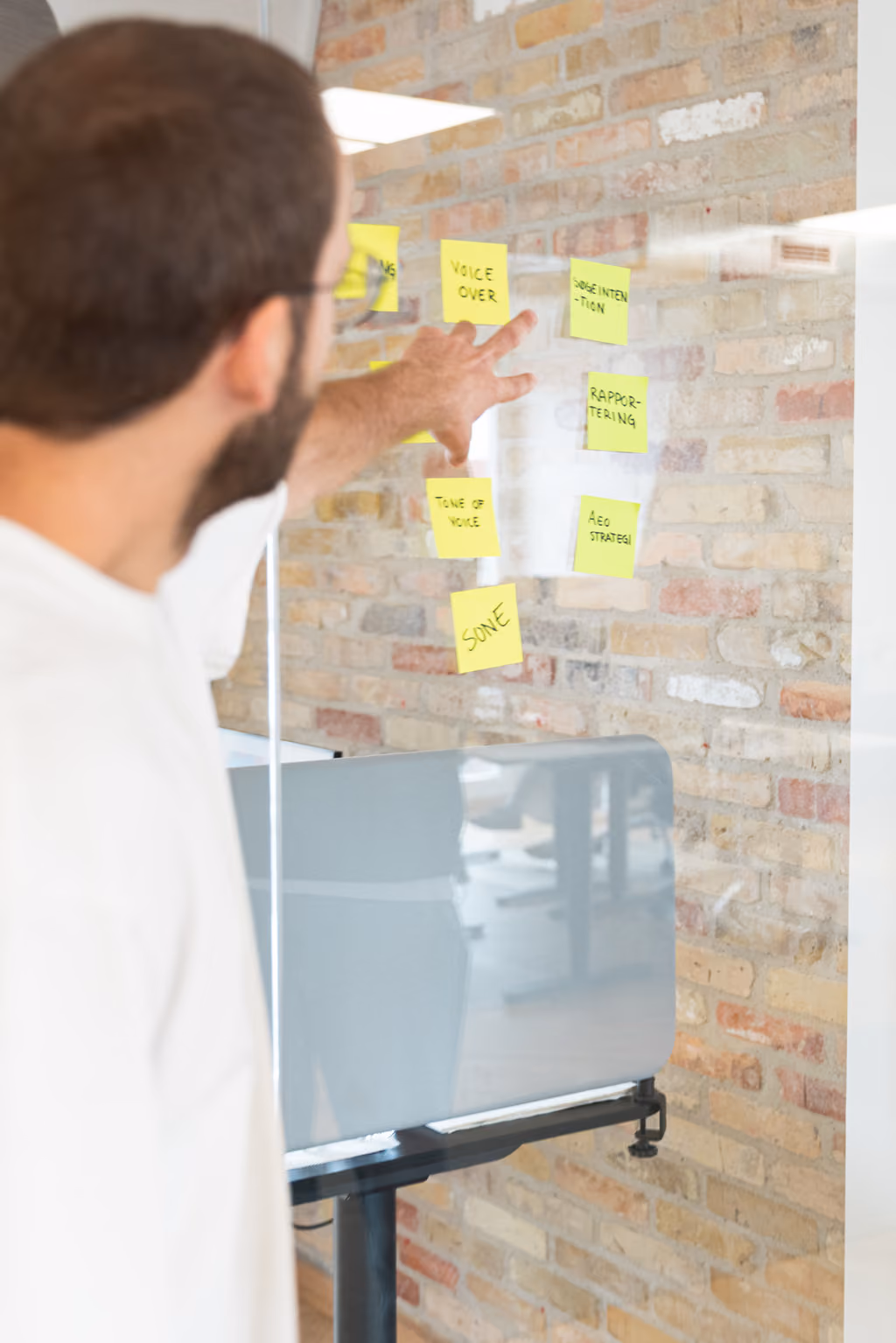 Mand peger på gule post-its med noter som 'voice over' på et glas i et kontormiljø.