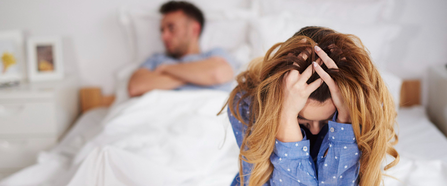 A couple sitting on the bed: the man with his arms crossed, the woman looking sad