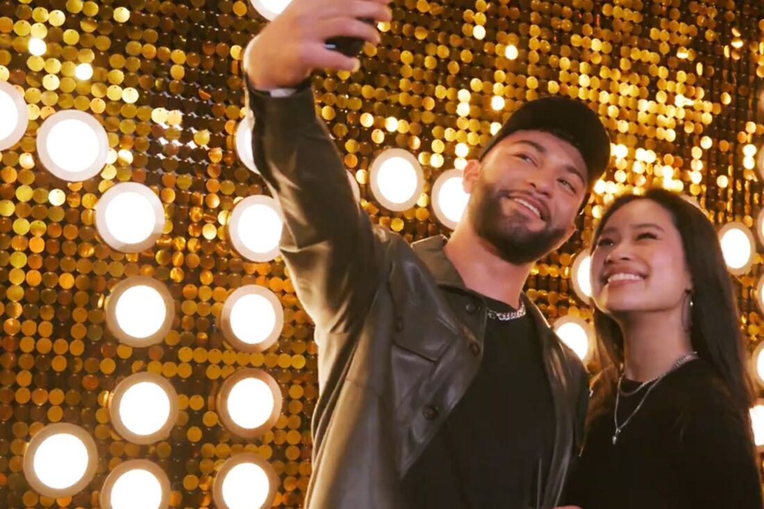 two people taking a selfie in front of a gold sequin wall