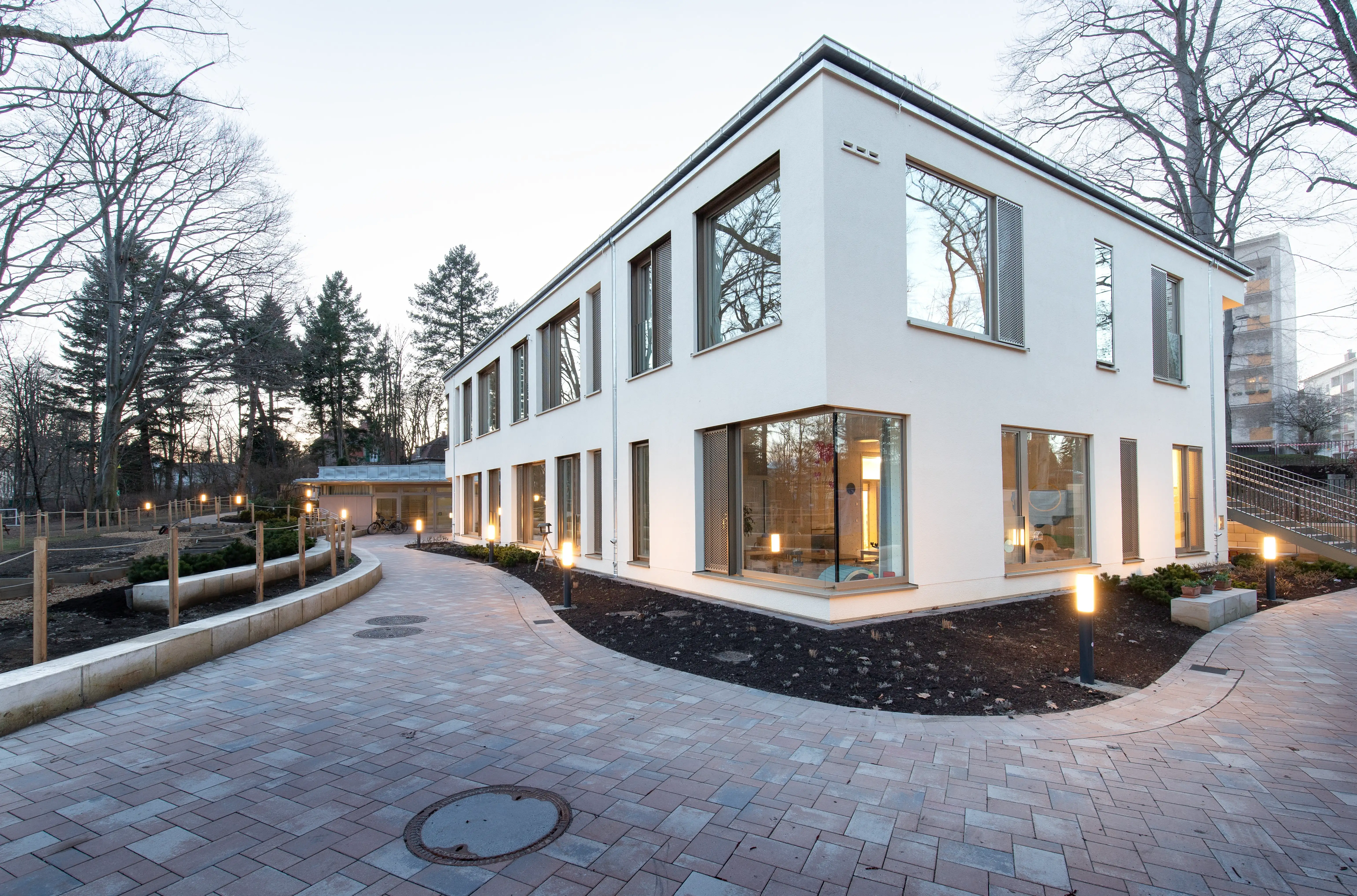 Modern zweistöckiges Kita-Gebäude mit großen Fenstern, beleuchteten Wegen und kahlen Bäumen im Hintergrund.
